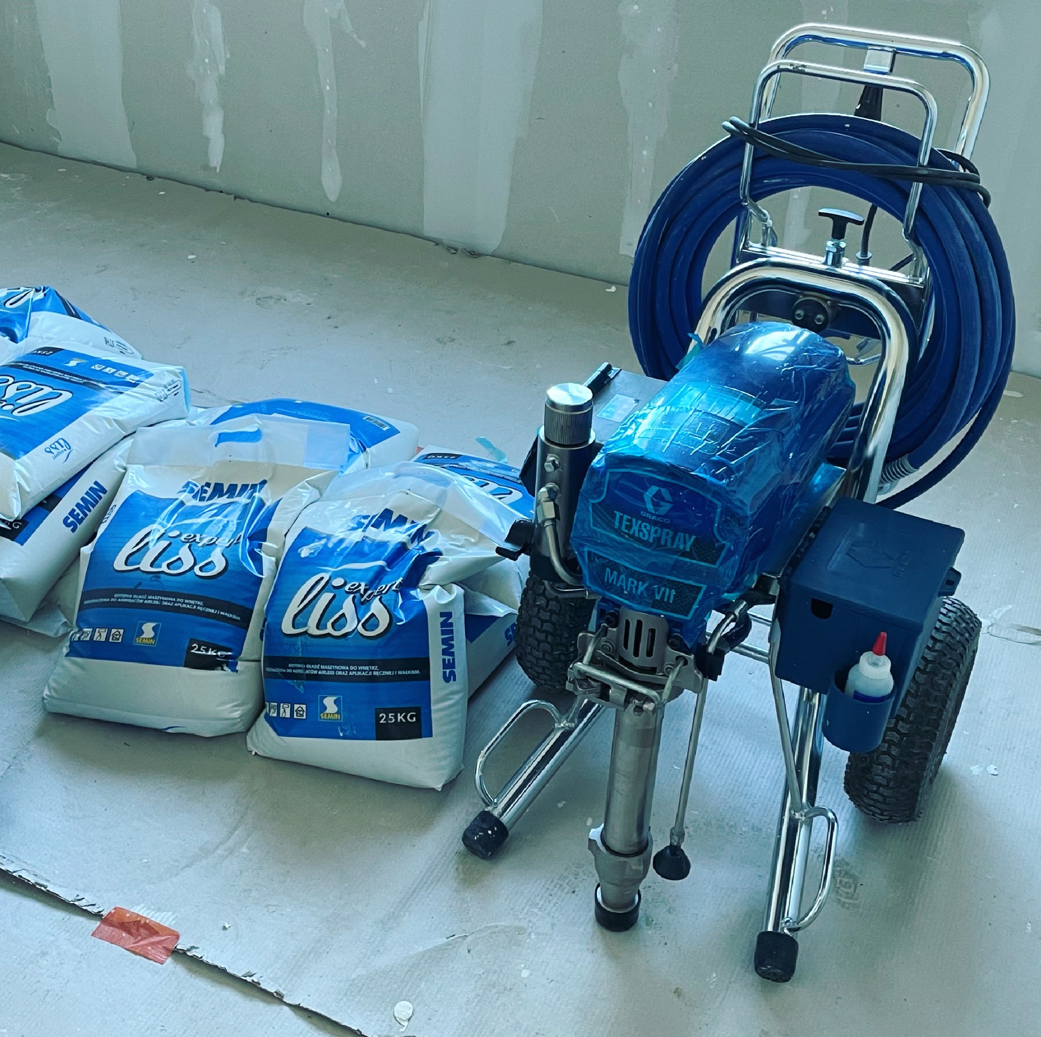 Agregat malarski Graco TexSpray Mark VII, owinięty folią ochronną, stoi obok stosu worków z gładzią szpachlową Semini Liss Expert 25kg, na surowym podłożu w trakcie prac wykończeniowych.