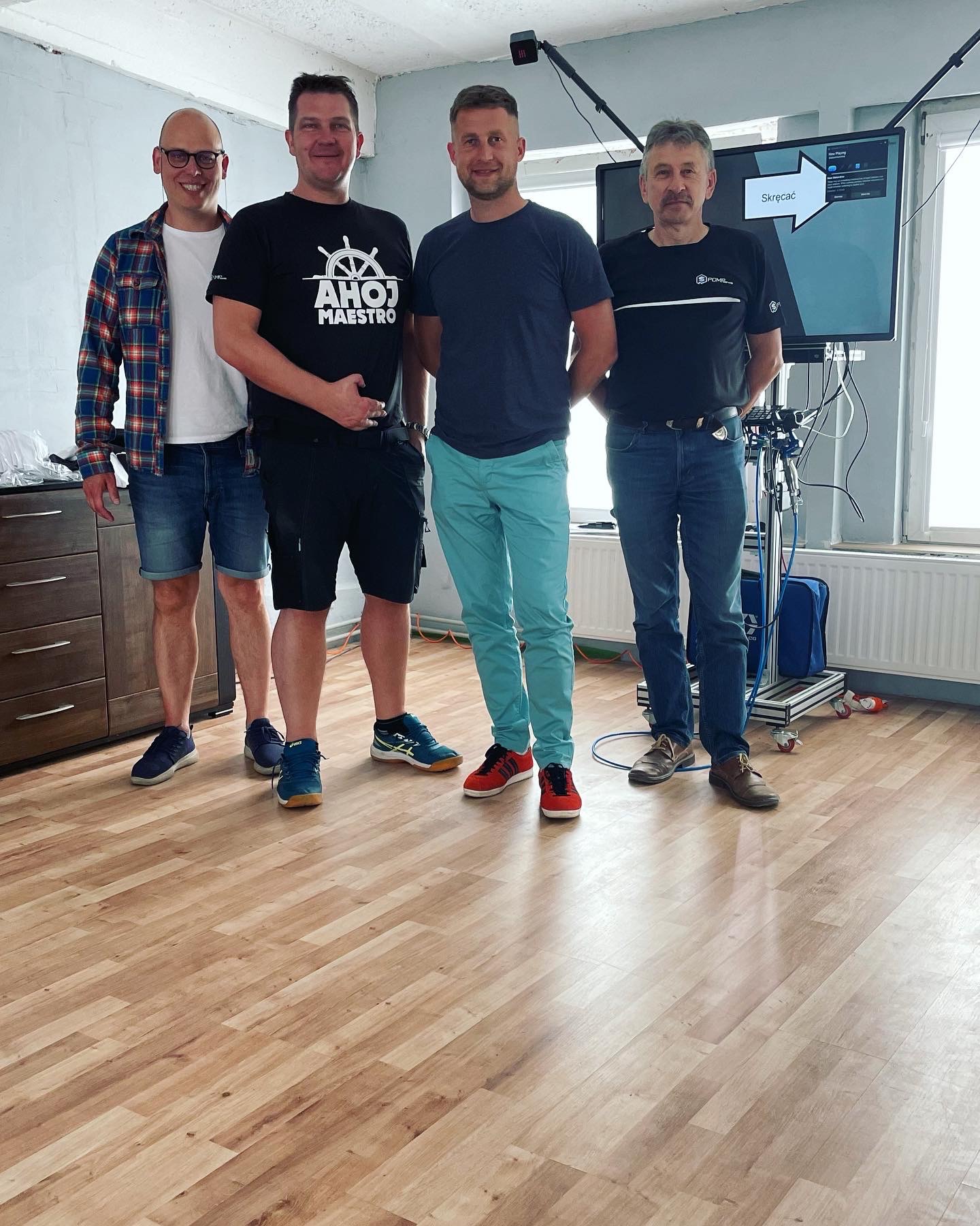 Czterech mężczyzn pozuje do zdjęcia w pomieszczeniu z drewnianą podłogą, widoczny sprzęt oświetleniowy i monitor z napisem 'Skręcać'.