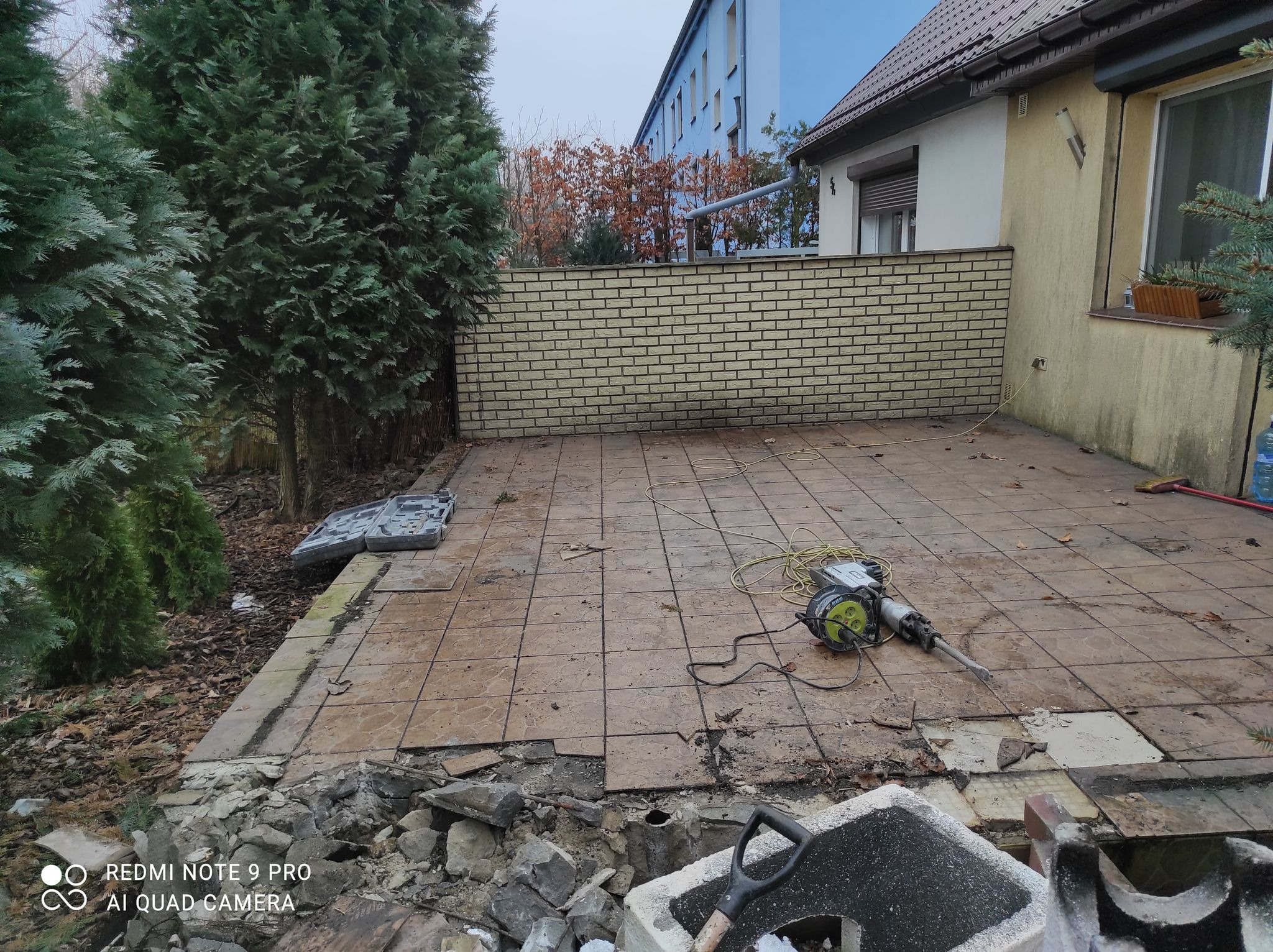 Demontaż starej, brązowej terakoty na tarasie przydomowym przy użyciu młota pneumatycznego; widoczne gruz, odspojone płytki i narzędzia.