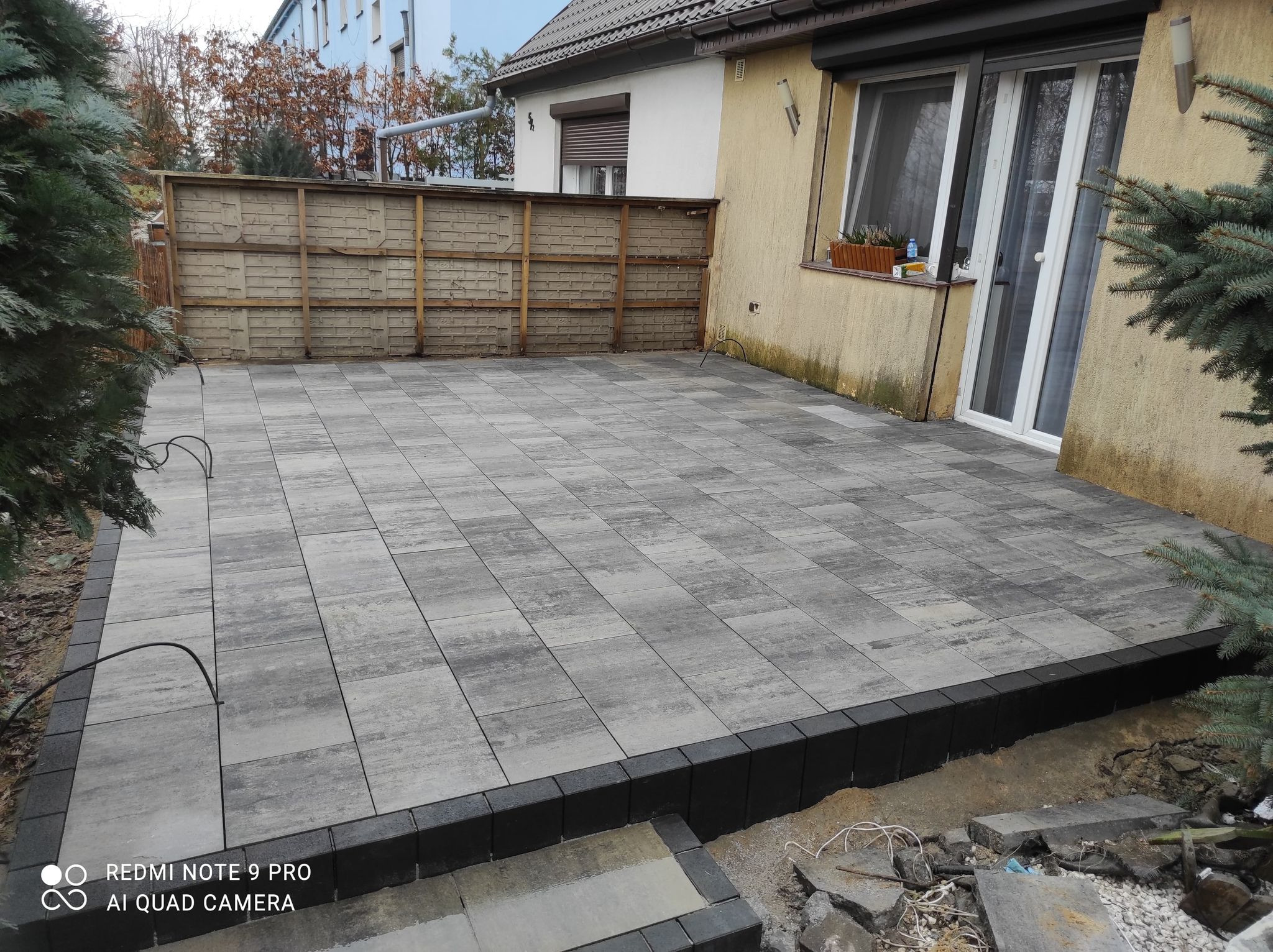 Ułożona szara kostka brukowa na podwórku z czarnym obramowaniem, widoczne elementy ogrodu i fragment domu z oknem i drzwiami balkonowymi.