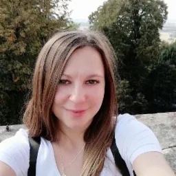 Kobieta z brązowymi włosami i srebrnym naszyjnikiem, uśmiechnięta, na tle drzew i krajobrazu z lekką mgłą w oddali.