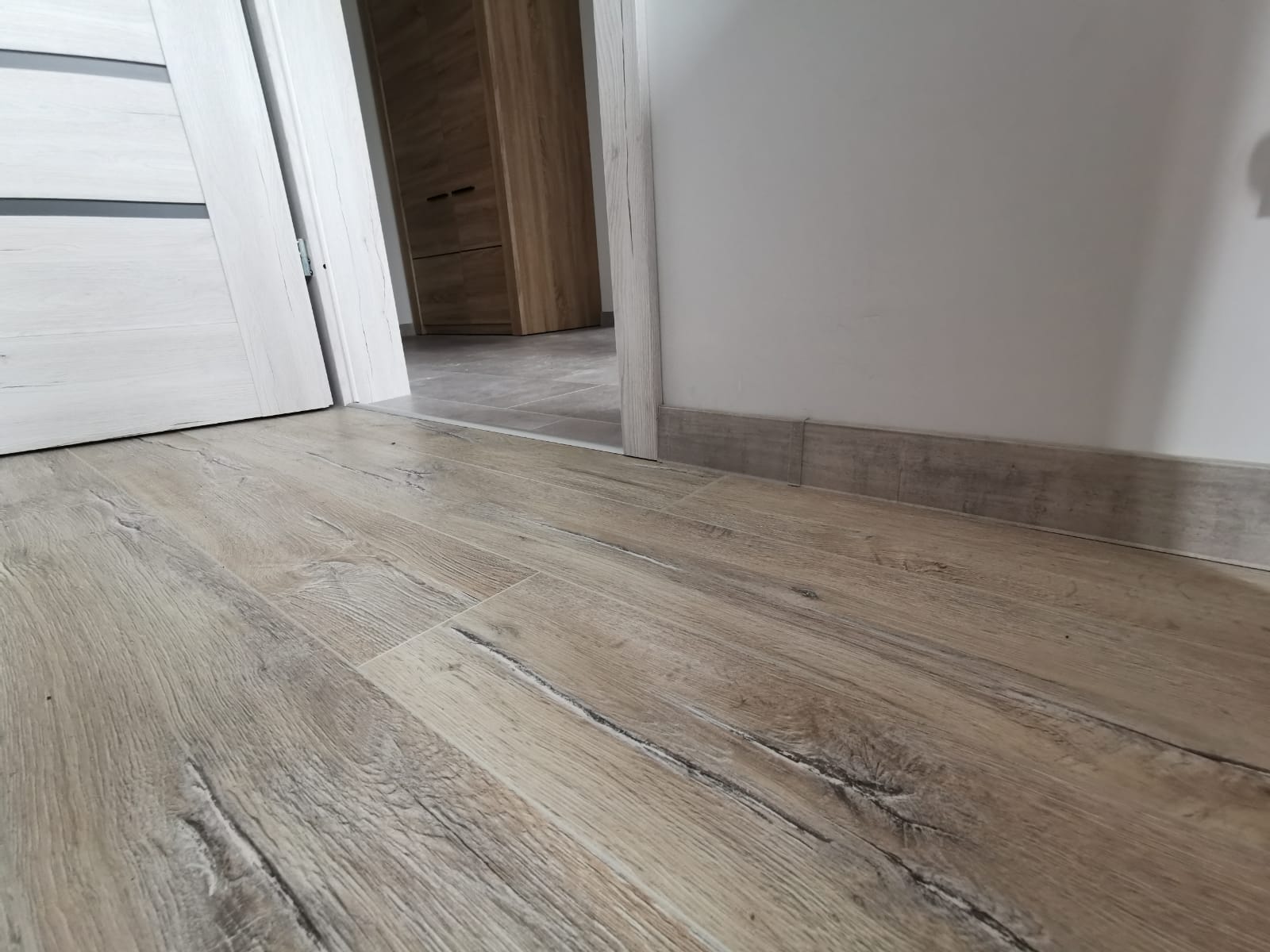 Ułożona podłoga z paneli imitujących drewno, widoczne listwy przypodłogowe i fragment drzwi wejściowych.