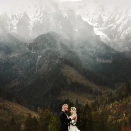 Para młoda w objęciach na tle mglistych gór i lasu, panna młoda w wianku z kwiatów.