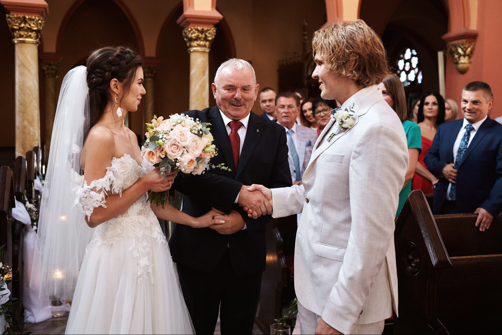 Uśmiechnięta panna młoda w białej sukni i welonie, ściska dłoń z uśmiechniętym panem młodym w jasnym garniturze, w tle goście i urzędnik w kościele.