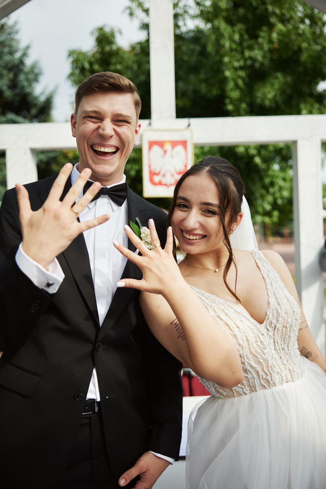 Uśmiechnięta para młoda prezentuje obrączki na tle białej konstrukcji i zieleni. Widoczny godło Polski. Panna młoda w sukni z aplikacjami, pan młody w czarnym smokingu.