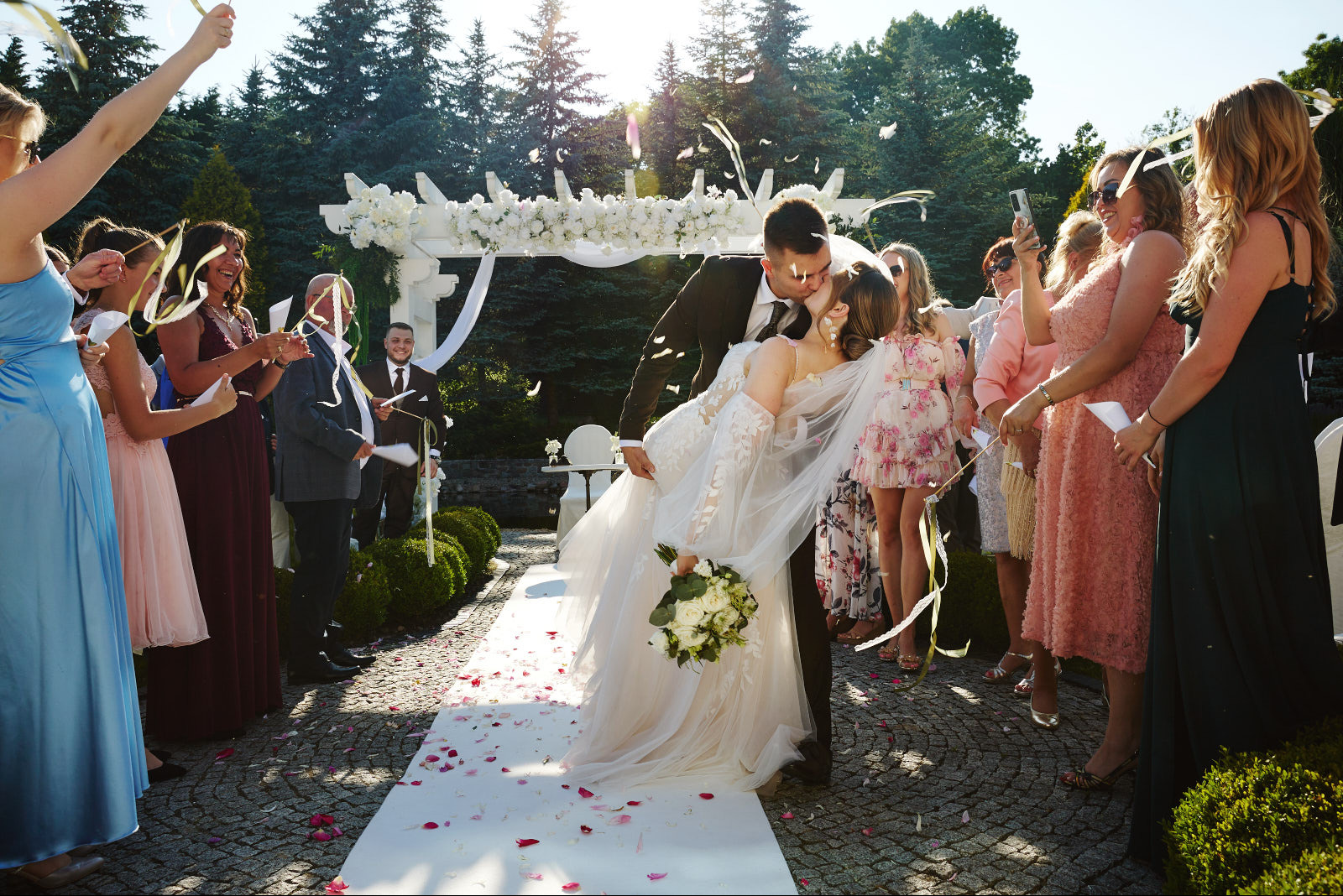 Pocałunek pary młodej na tle białej pergoli, goście rzucają konfetti. Pan młody trzyma pannę młodą w ramionach. Ujęcie w słoneczny dzień, zieleń w tle.