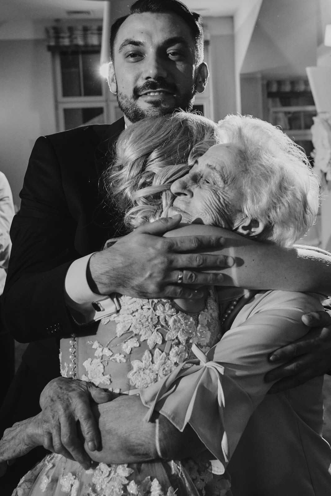 Czarno-białe zdjęcie wzruszającego uścisku panny młodej i starszej kobiety, otulonych ramionami mężczyzny w garniturze, uchwycone podczas uroczystości ślubnej.