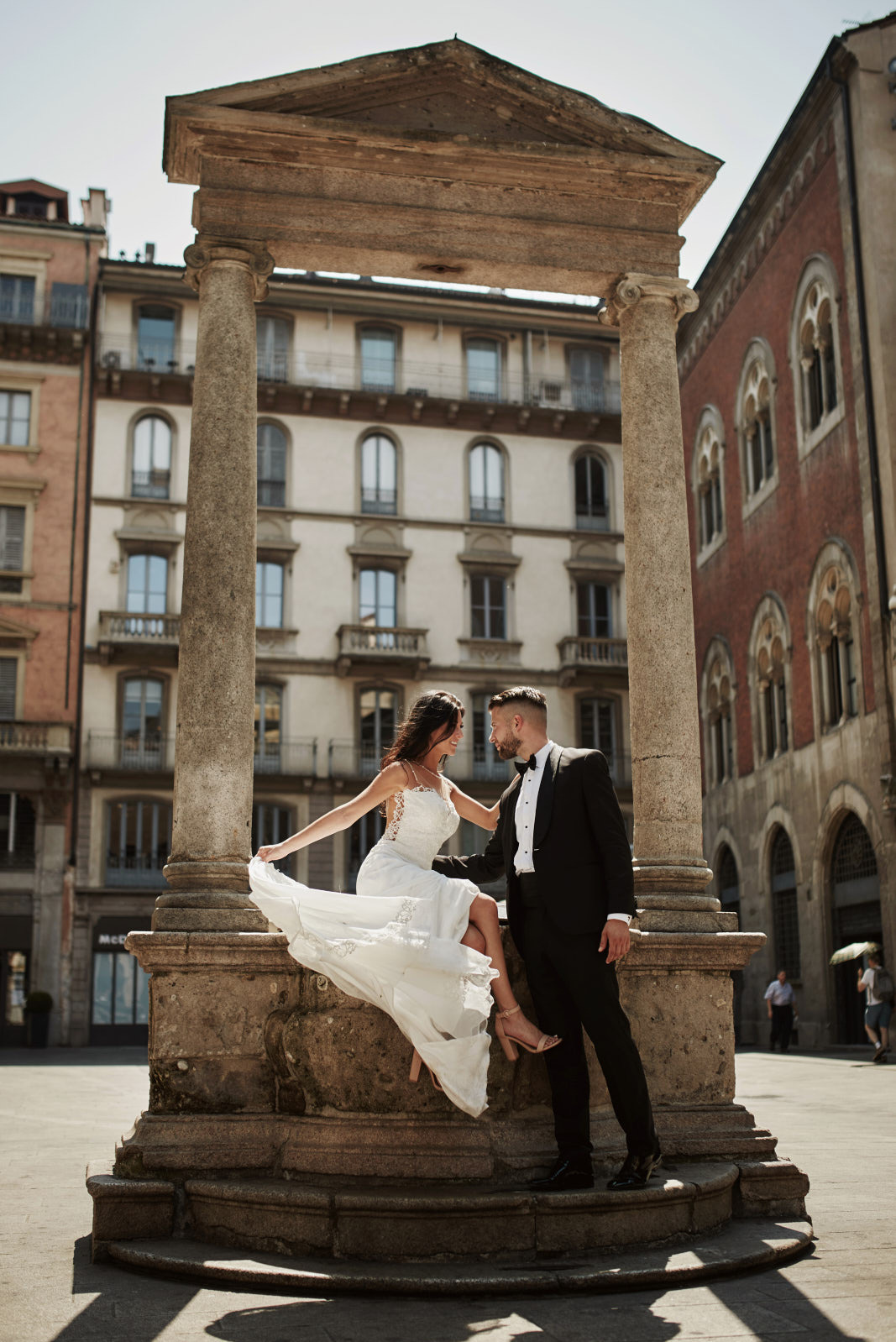 Para młoda w eleganckich strojach pozuje na tle kolumn i miejskiej zabudowy. Panna młoda siedzi, trzymając suknię, pan młody stoi obok. Ujęcie w słoneczny dzień.