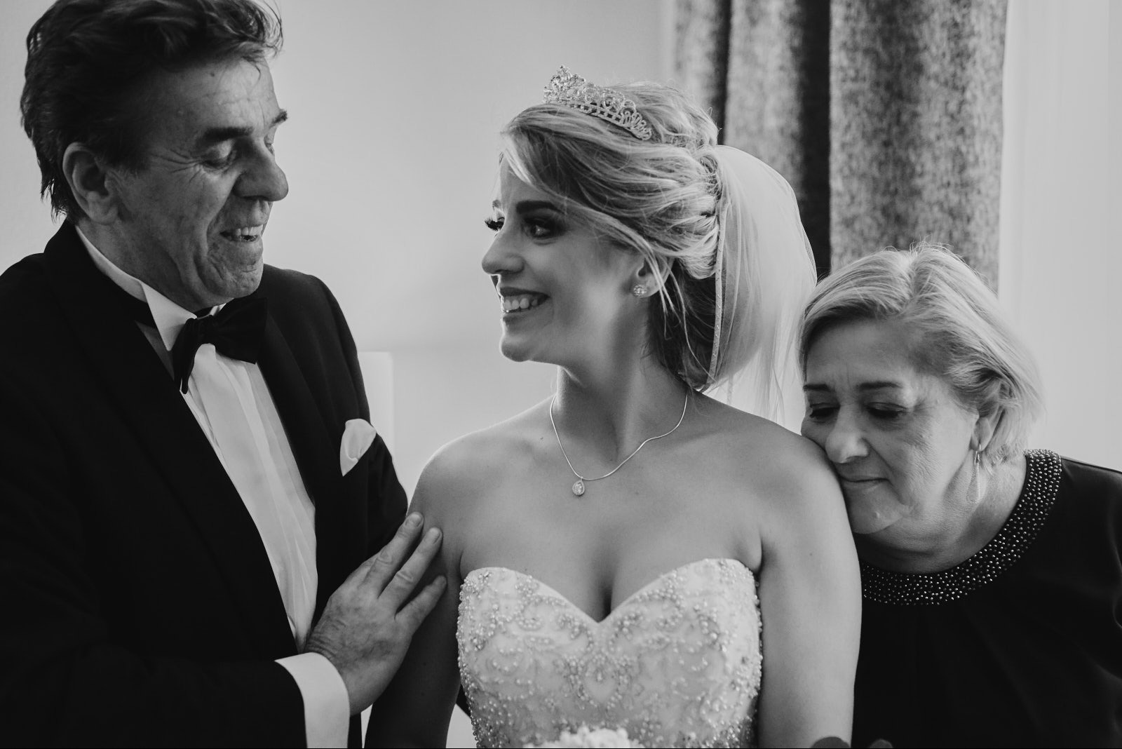 Czarno-białe zdjęcie uśmiechniętej panny młodej w sukni ślubnej z tiarą, otoczonej przez ojca w smokingu i matkę, w dniu ślubu, pełne emocji i wzruszeń.