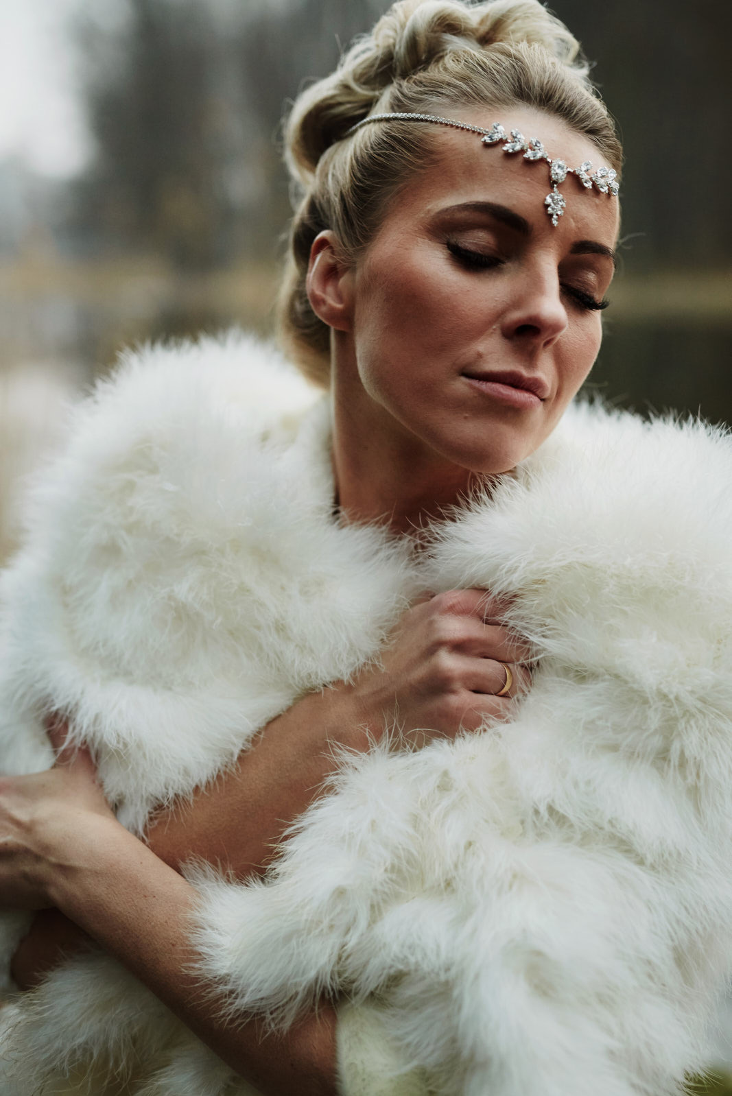 Portret kobiety w białym futrze i ozdobnej opasce na głowie, z zamkniętymi oczami, w naturalnym oświetleniu, na rozmytym tle.
