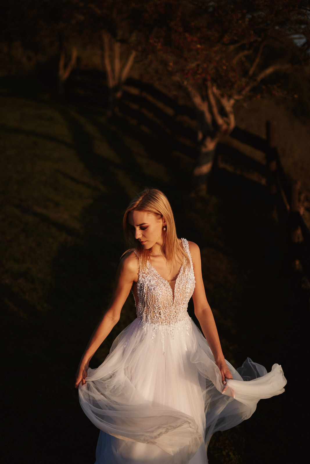 Elegancka blondynka w koronkowej sukni ślubnej, delikatnie trzymająca materiał. Ujęcie z góry, ciepłe światło zachodzącego słońca, tło z zielenią i drewnianym płotem.