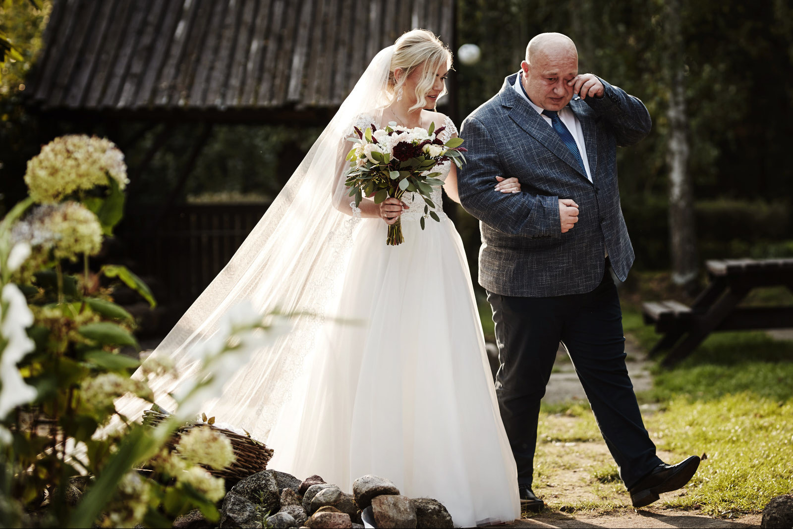 Panna młoda w białej sukni i welonie, trzyma bukiet, prowadzona do ołtarza przez wzruszonego ojca w szarym garniturze na tle zieleni i drewnianej altany.