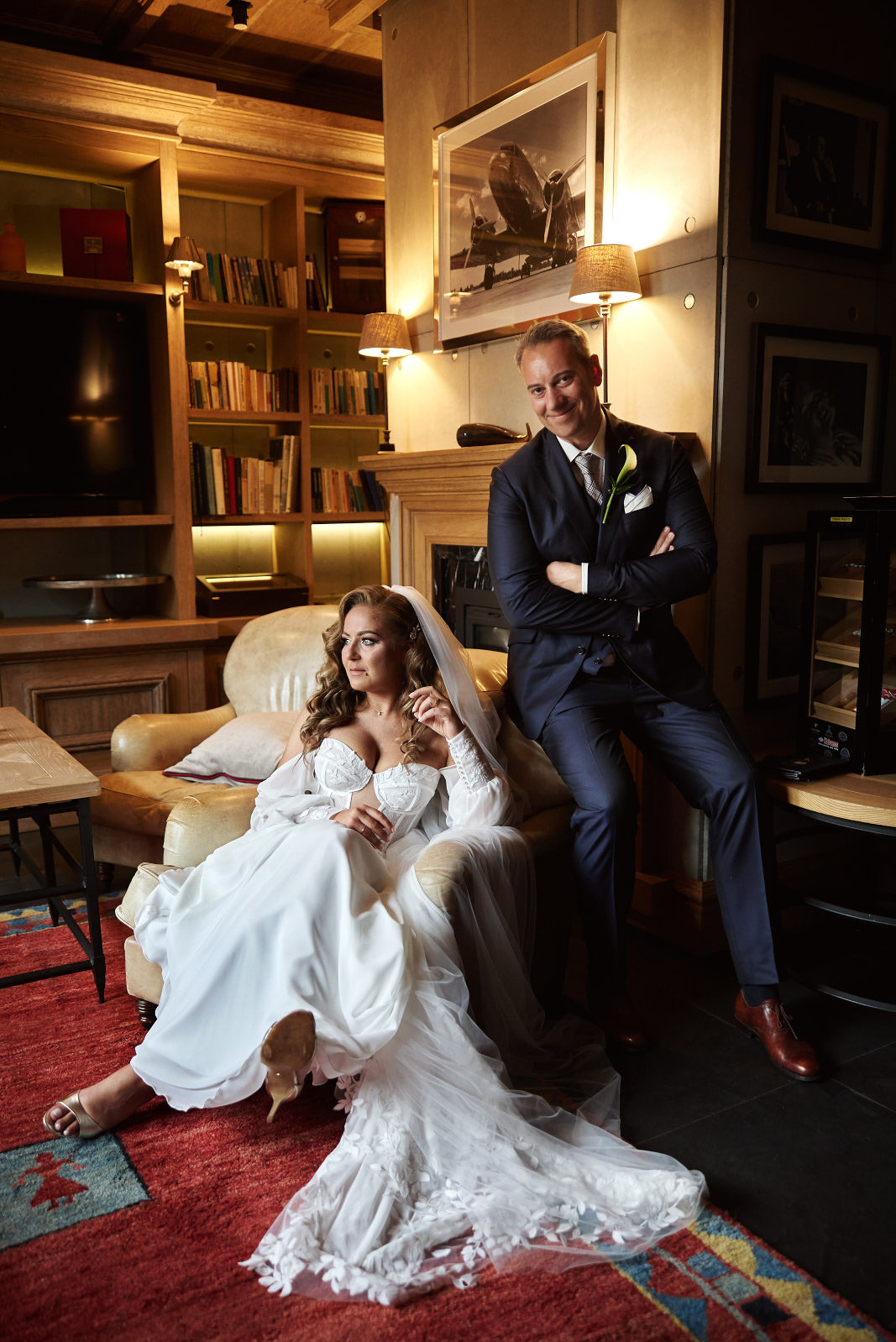 Elegancka sesja ślubna w stylowym wnętrzu: panna młoda w koronkowej sukni siedzi w fotelu, obok stoi pan młody w granatowym garniturze. W tle biblioteczka i kominek.