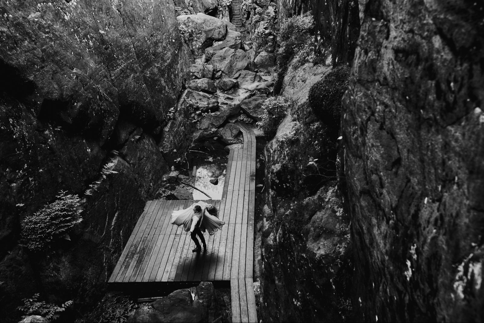 Czarno-białe zdjęcie panny młodej na drewnianym pomoście w wąwozie skalnym. Ujęcie z góry podkreśla surowość otoczenia i delikatność sukni.