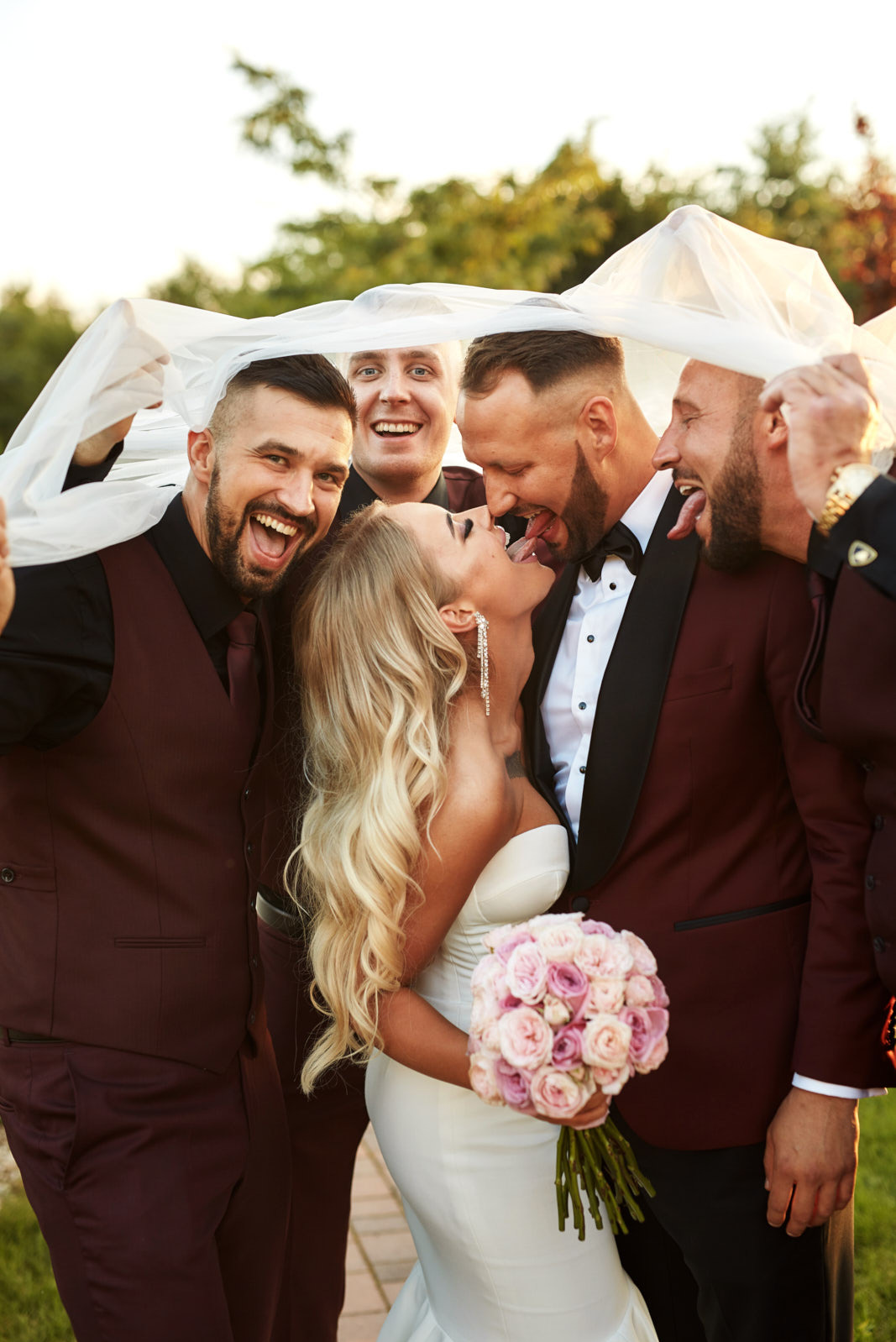 Radosna para młoda z drużbami pod welonem, wyciągają języki w żartobliwym geście. Bukiet różowych róż, bordowe garnitury, uśmiechy i naturalne światło.