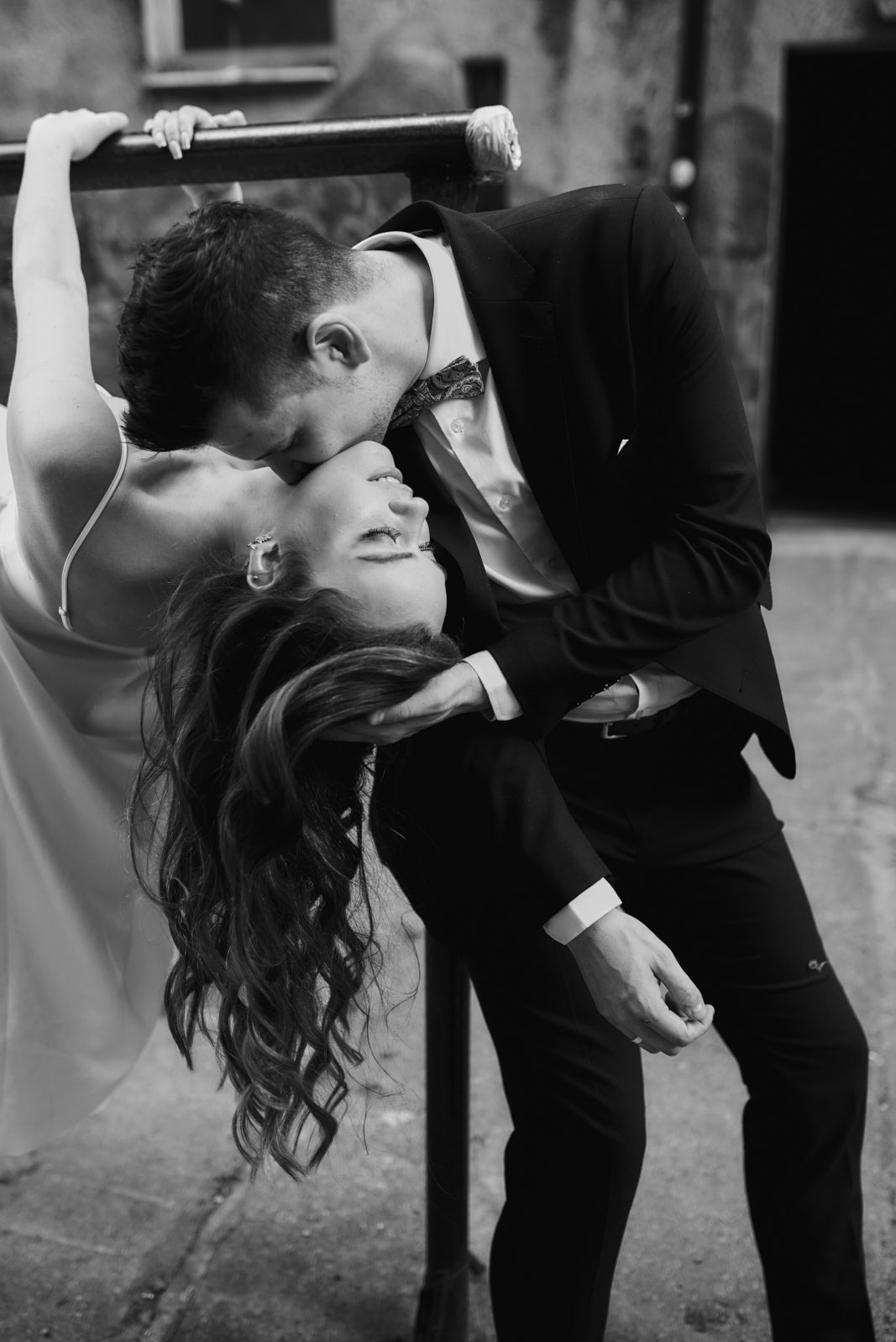 Czarno-białe zdjęcie pary młodej w tańcu: on w smokingu całuje ją w czoło, ona odchylona do tyłu, trzyma się barierki. Ujęcie z dołu, podkreślające dynamikę i emocje.
