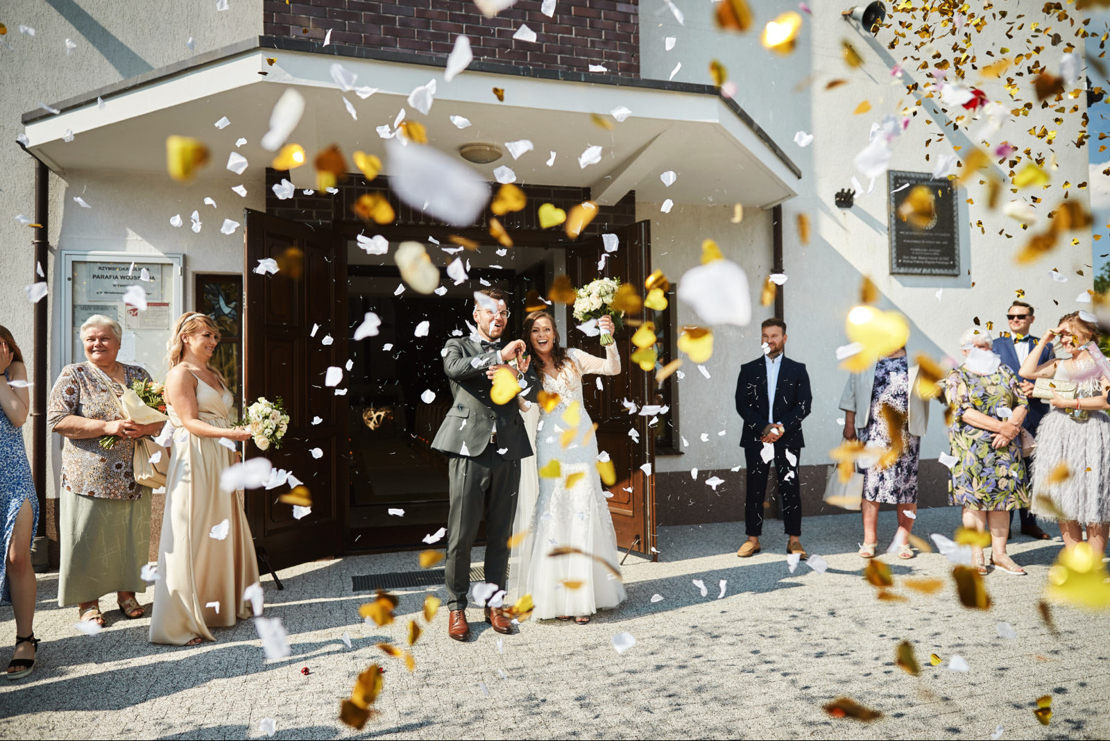 Uśmiechnięta para młoda wychodzi z kościoła, obsypywana konfetti w kształcie serc i płatków. W tle goście weselni i jasna fasada budynku.