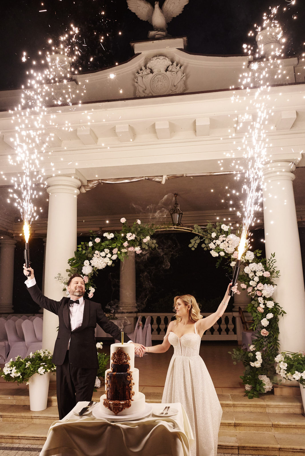 Uśmiechnięta para młoda z fontannami iskier przy tort weselnym na tle kolumnady z kwiatową dekoracją. Pan młody w smokingu, panna młoda w sukni z brokatem.