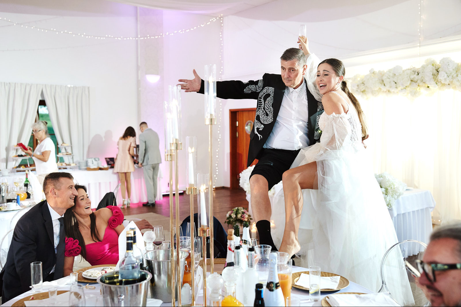 Rozbawiona para młoda w nietypowych strojach tańczy na stole podczas wesela. W tle goście, dekoracje kwiatowe i świecące girlandy. Ujęcie z perspektywy gościa.