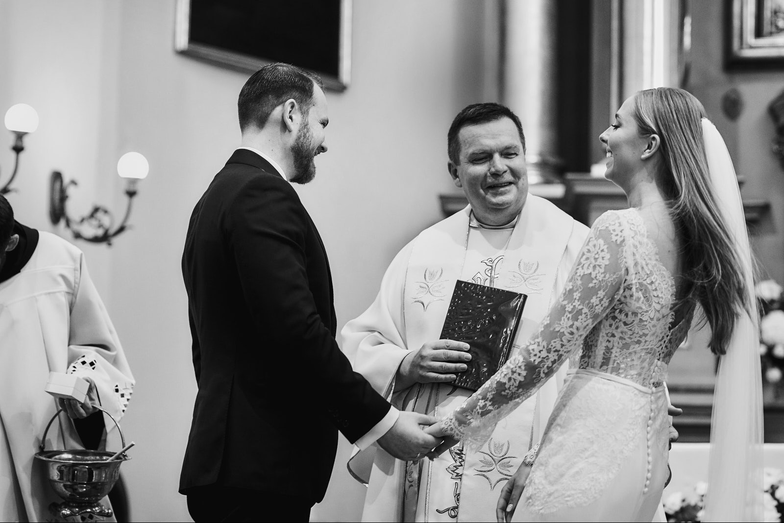 Czarno-białe zdjęcie z ceremonii ślubnej: uśmiechnięta para trzyma się za ręce przed duchownym z księgą. W tle widoczne elementy wystroju kościoła.