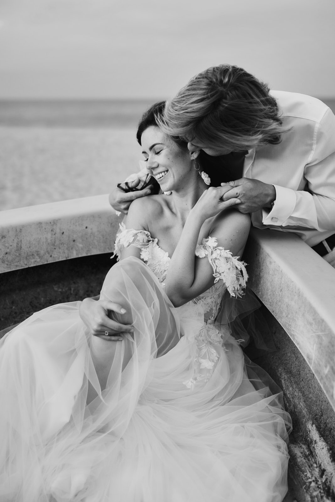 Czarno-białe zdjęcie uśmiechniętej panny młodej w koronkowej sukni, obejmowanej przez pana młodego na tle plaży. Romantyczny kadr z sesji narzeczeńskiej.