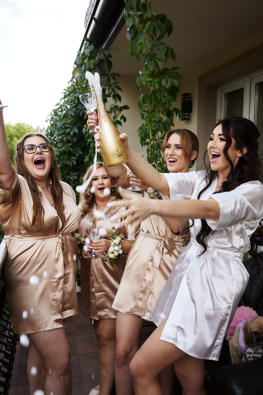 Radosne kobiety w szlafrokach świętują, otwierając szampana. W tle zielona roślinność i budynek. Ujęcie z dołu podkreśla dynamikę i energię sceny.