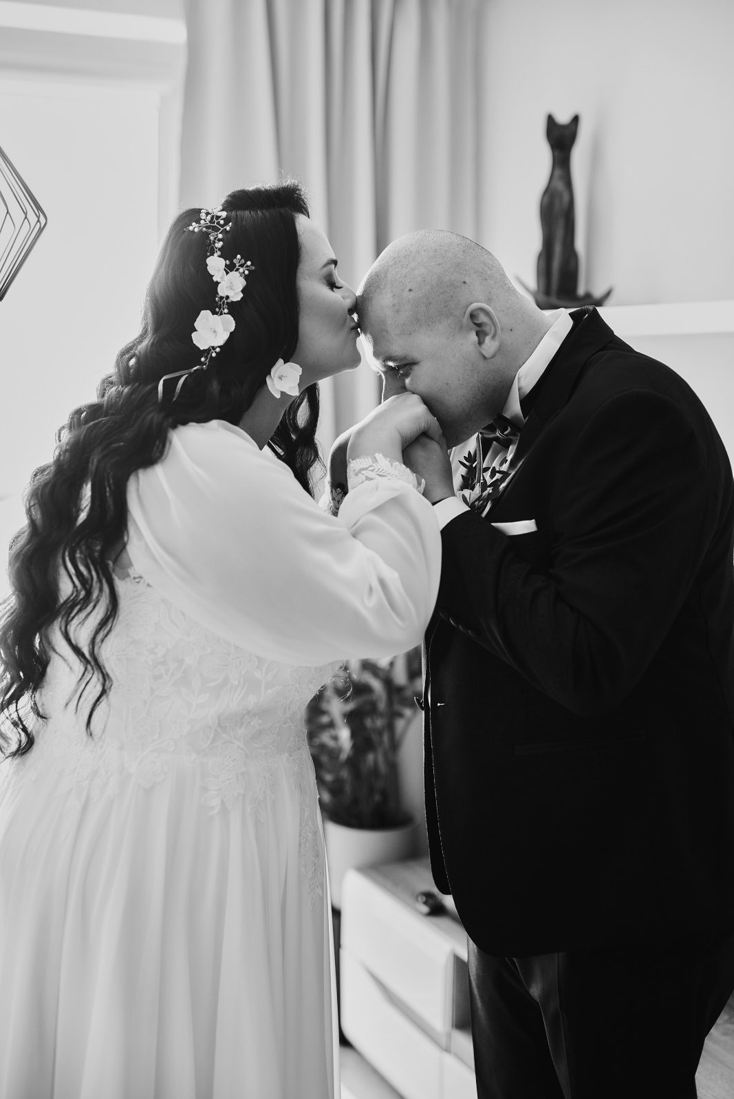 Czarno-białe zdjęcie panny młodej w kwiecistej ozdobie włosów, całującej dłoń pana młodego w eleganckim smokingu, na tle jasnego wnętrza z dekoracyjną figurką kota.