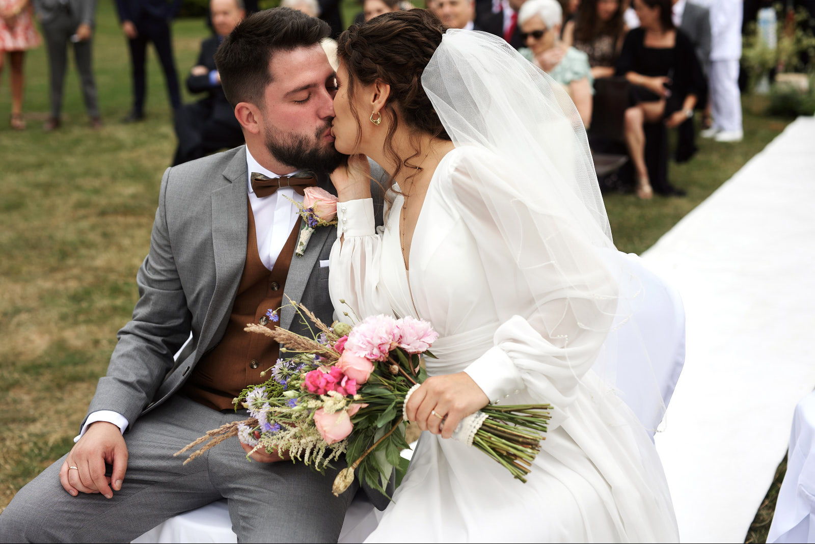 Pocałunek nowożeńców siedzących na tle gości weselnych. Panna młoda w białej sukni z bukietem, pan młody w szarym garniturze z brązową muchą.