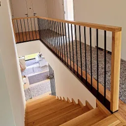 Schody drewniane z jasnego drewna z czarną balustradą, widok z góry, w tle drzwi i fragment podłogi z lastryko.