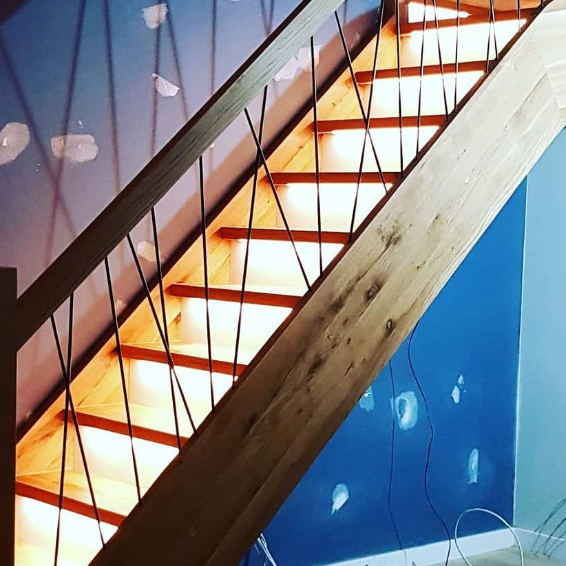 Nowoczesne schody drewniane z czarnymi, krzyżującymi się balustradami, podświetlane stopnie, niebieska ściana w trakcie wykończenia.