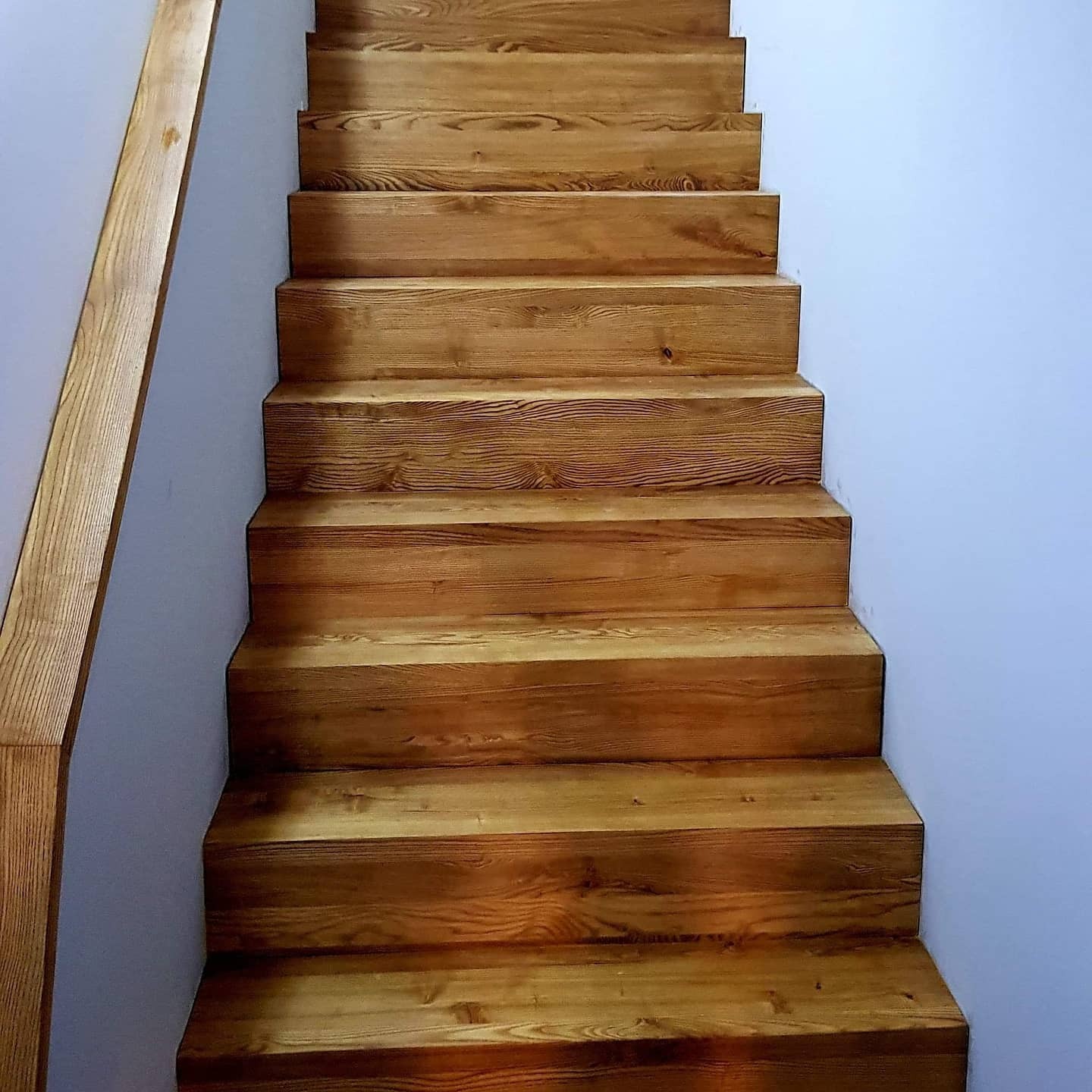 Proste schody z jasnego drewna z widocznym rysunkiem słojów, zamontowane przy białej ścianie, z widoczną częścią drewnianej balustrady po lewej stronie.