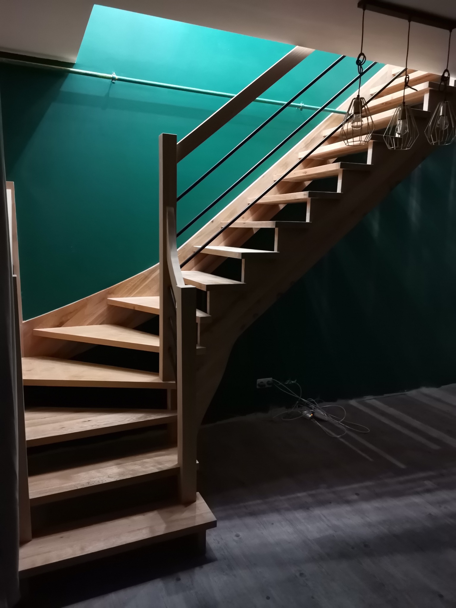 Jasne, drewniane schody z czarnymi, metalowymi barierkami na tle turkusowej ściany, oświetlone wiszącymi lampami drucianymi
