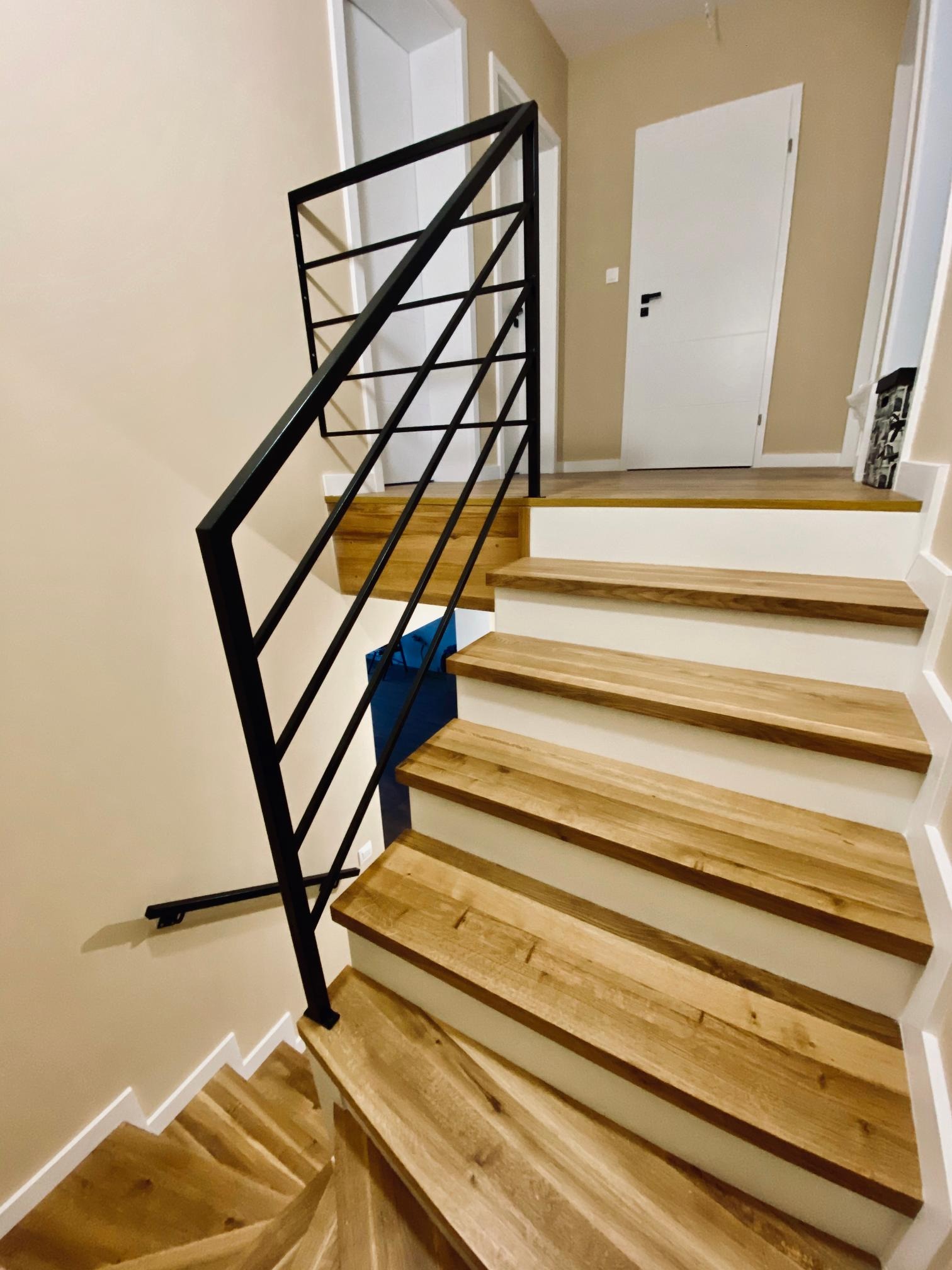 Nowoczesne schody z jasnego drewna z czarną metalową balustradą, widok z dołu, białe podstopnice, beżowe ściany, białe drzwi w tle.