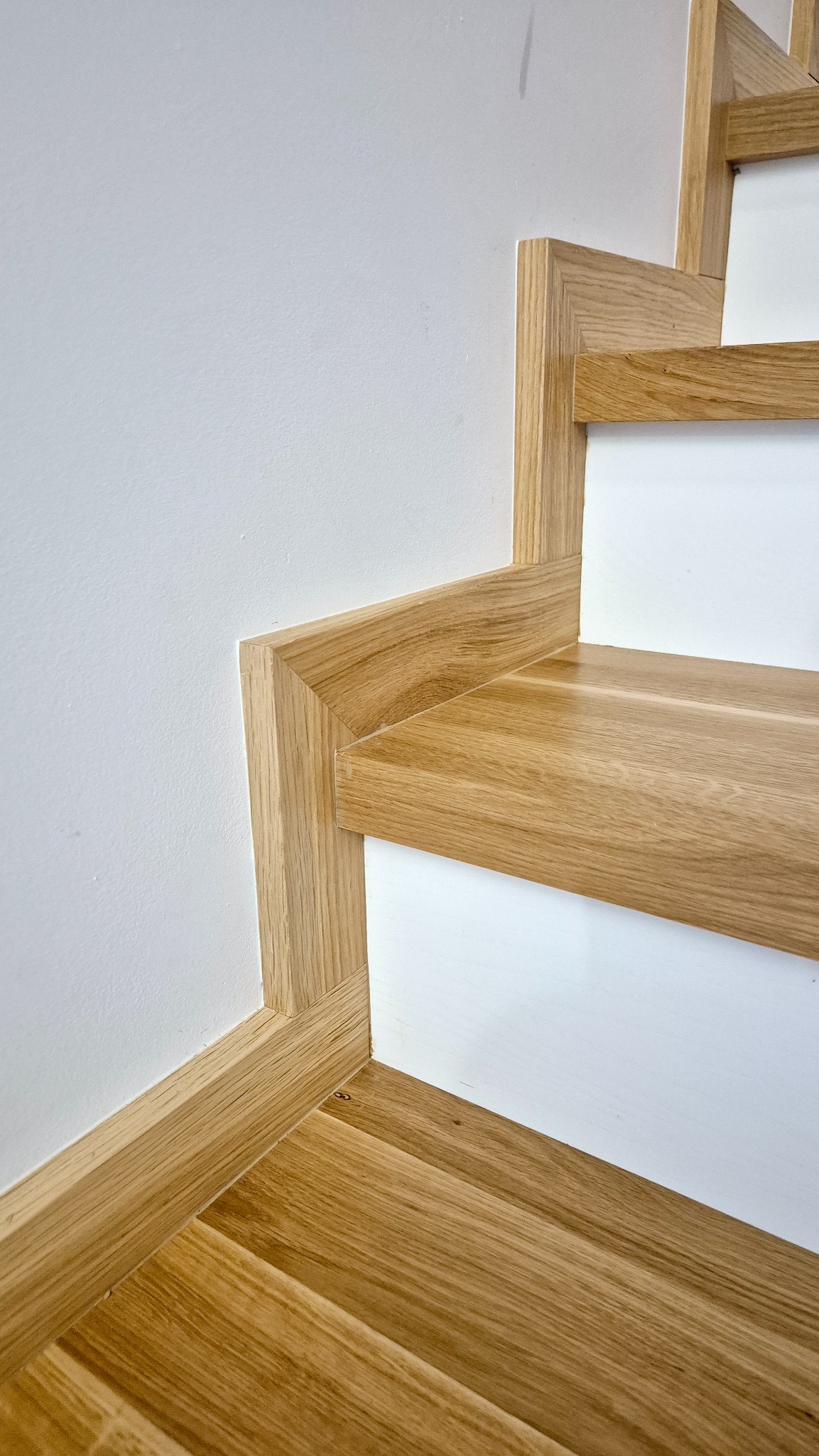 Fragment drewnianych schodów z białymi podstopnicami, widoczne połączenie z listwą przypodłogową i ścianą, jasne drewno o wyraźnym usłojeniu.