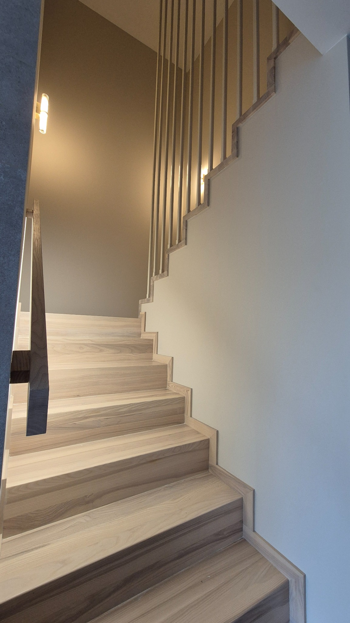 Jasne, minimalistyczne schody z drewna jesionowego z prostą balustradą, oświetlone dwoma kinkietami ściennymi.