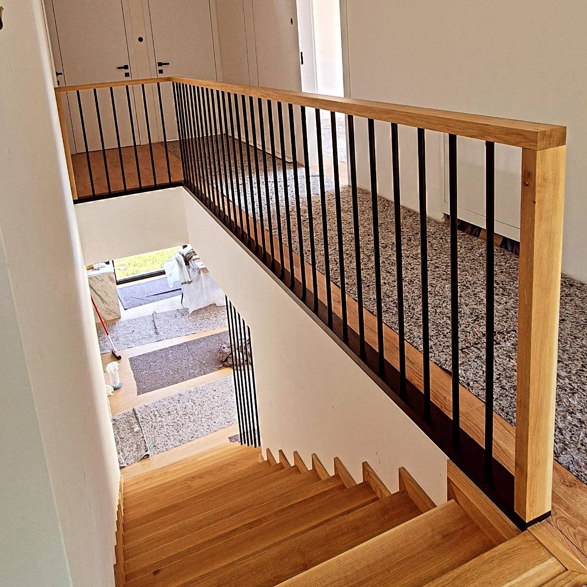 Schody drewniane z jasnego drewna z czarną balustradą, widok z góry, w tle drzwi i fragment podłogi z lastryko.