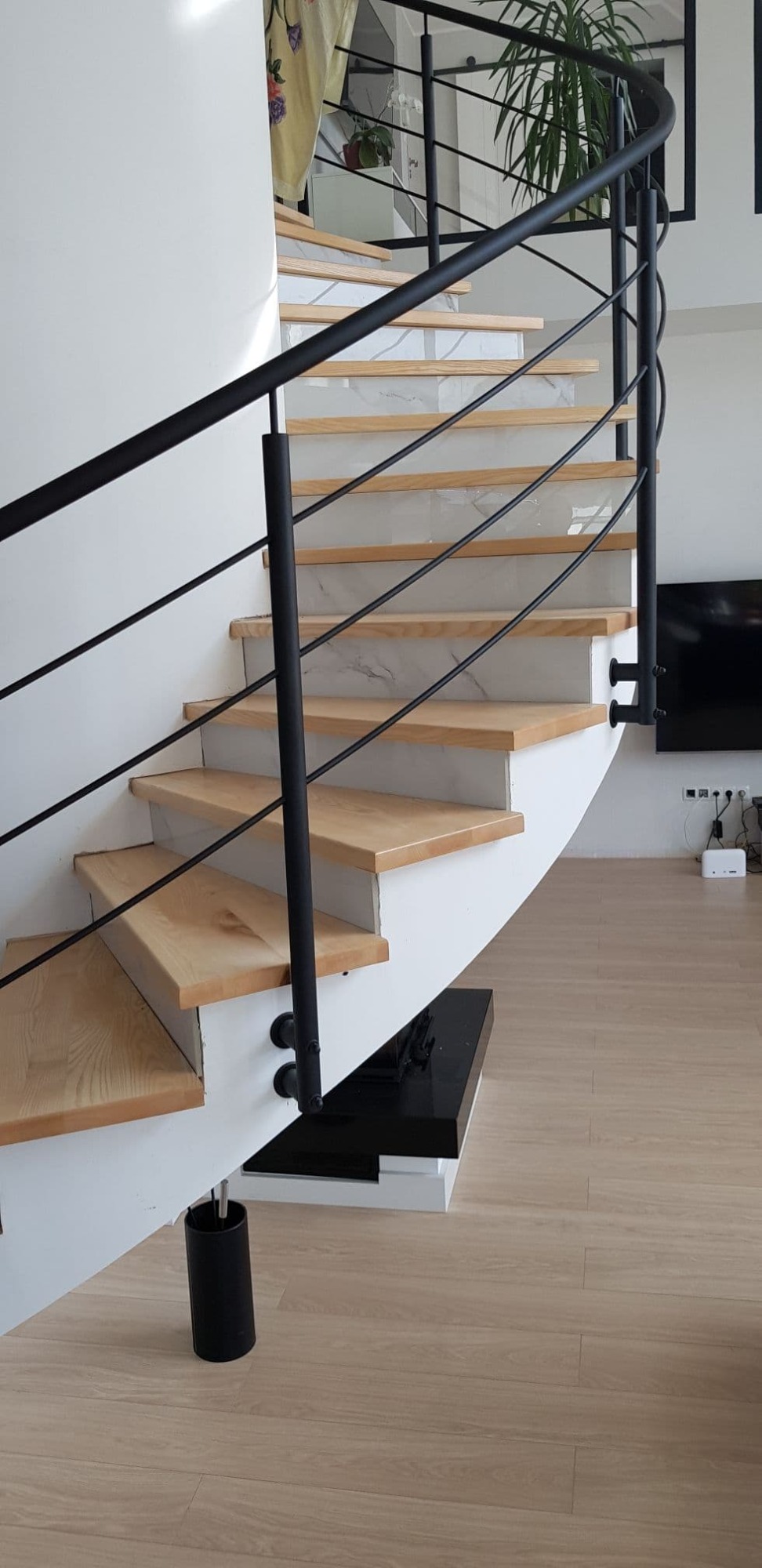 Spiralne schody z jasnego drewna z czarną metalową balustradą, stopnie wykończone białymi płytkami imitującymi marmur, widok od dołu.