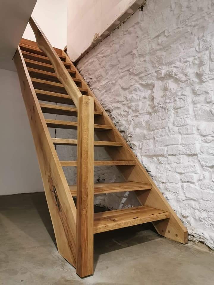 Drewniane schody z naturalnego drewna, widoczne sęki i usłojenie, zamontowane przy białej, surowej ścianie z kamienia.