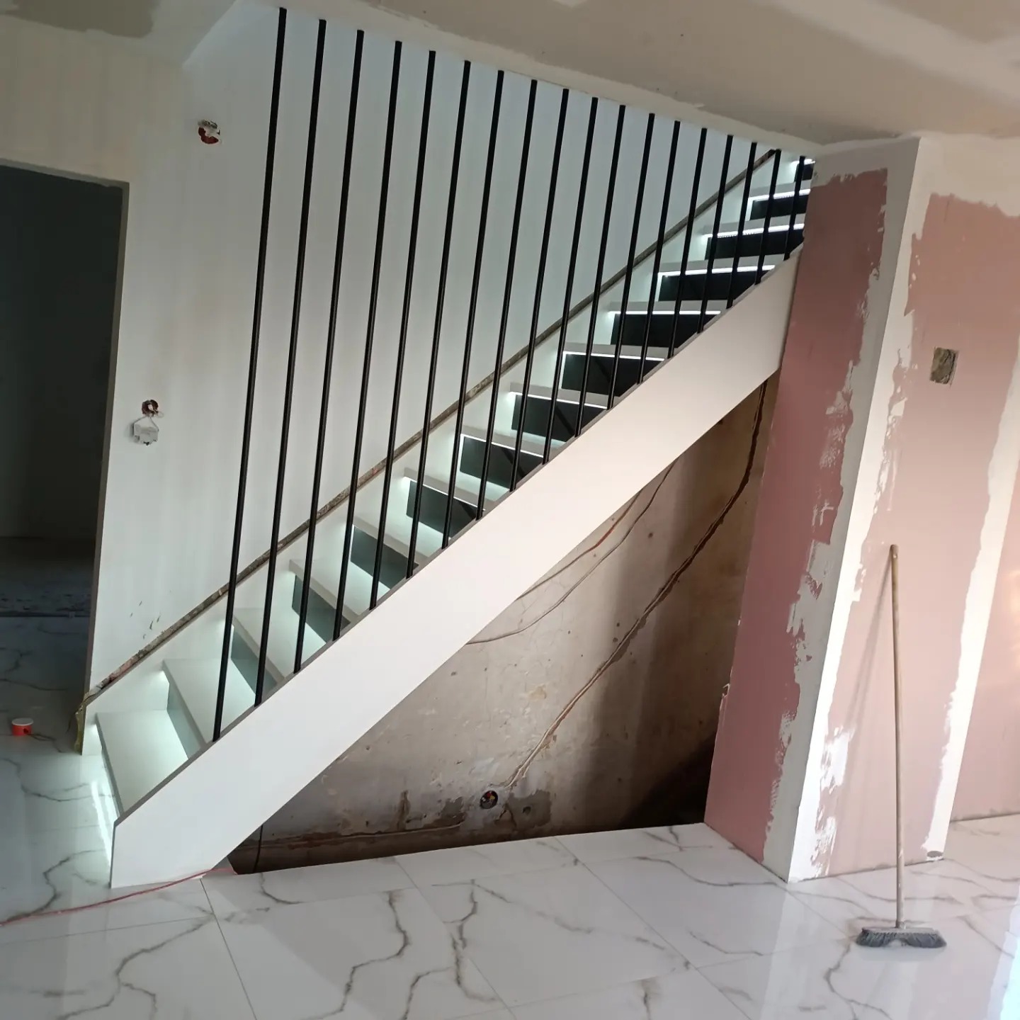 Schody drewniane z czarnymi balustradami w trakcie montażu, widoczne niedokończone ściany i podłoga z białego marmuru.