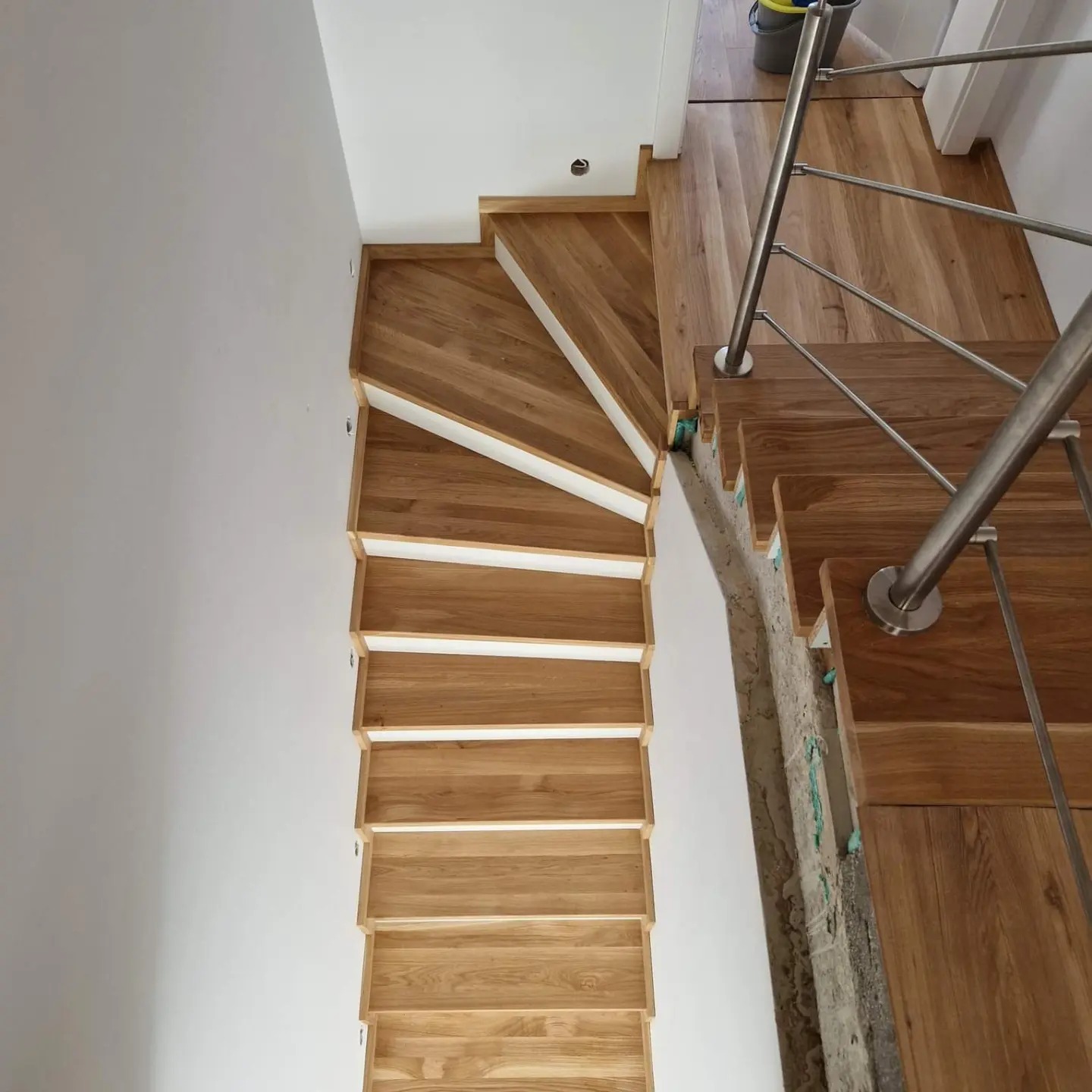 Widok z góry na jasne, drewniane schody z białymi podstopnicami, prowadzące w górę, z elementami metalowej balustrady i fragmentem surowej ściany obok stopni.