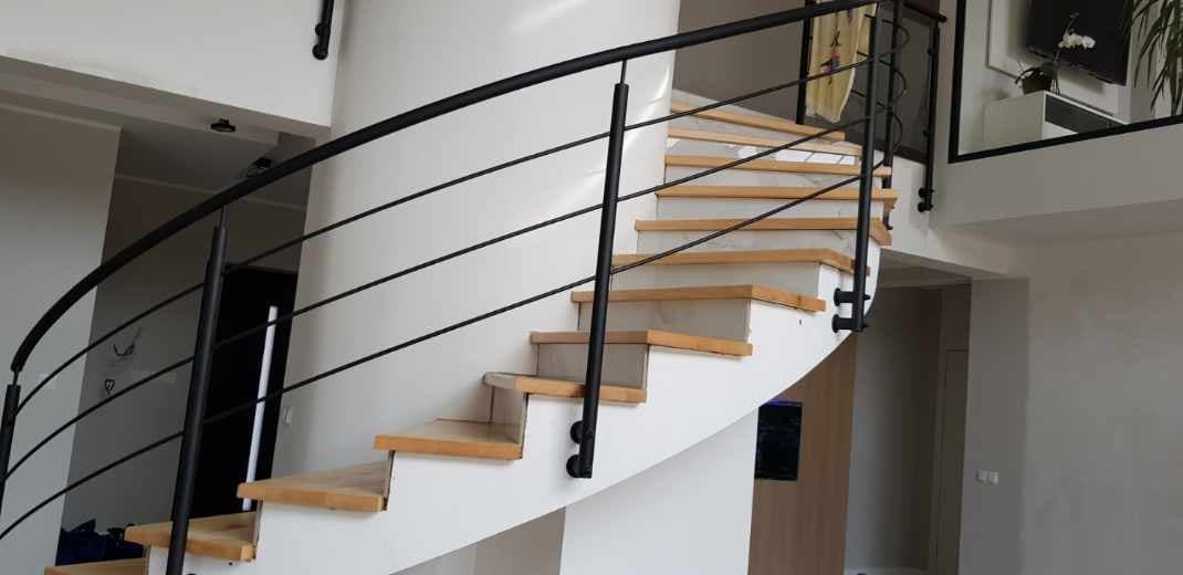 Spiralne schody z drewnianymi stopniami i czarną metalową balustradą, umieszczone przy białej, zakrzywionej ścianie w jasnym wnętrzu.