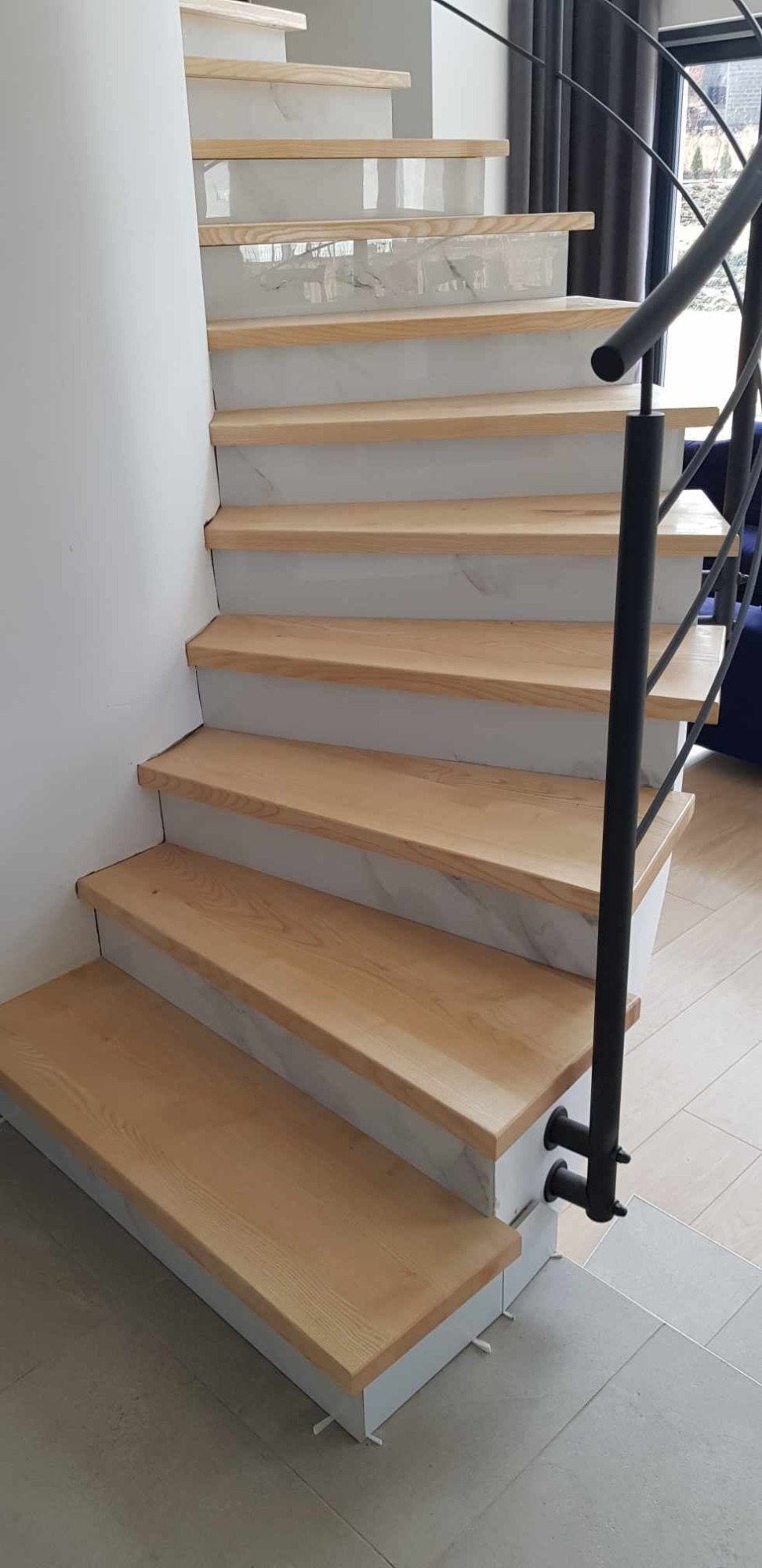 Jasne, drewniane stopnie schodów z marmurowymi podstopnicami i czarną, metalową balustradą, widok z dołu na pierwsze stopnie.