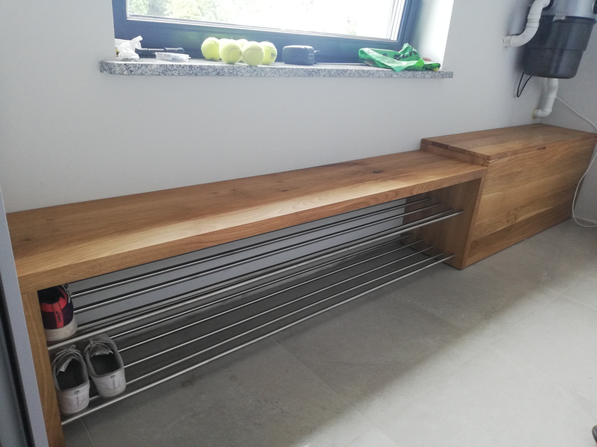 Drewniana ławka z półkami na buty wykonana z jasnego drewna, z widocznymi słojami, umieszczona pod oknem z piłkami tenisowymi na parapecie, na podłodze z szarych płytek.