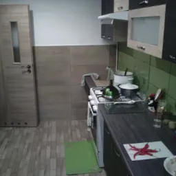 Kuchnia w Zagorzycach po remoncie: widok na nową zabudowę kuchenną z ciemnymi szafkami i zielonymi płytkami, drewnopodobne panele podłogowe, drzwi z przeszkleniem oraz detale jak czajnik...