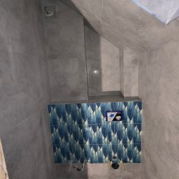 MK Remonty - Wykończenia - Ściana łazienki wyłożona szarymi płytkami imitującymi beton, z fragmentem dekoracyjnej ściany z niebieskimi płytkami w motywy piór, widoczne otwory na instalacje sanitarne i zamontowana podtynkowa...