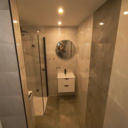 MK Remonty - Wykończenia - Nowoczesna łazienka z kabiną prysznicową typu walk-in, umywalką nablatową z szafką wiszącą i dużym okrągłym lustrem, wykończona płytkami w odcieniach beżu i szarości z widocznym odbiciem fotografa...