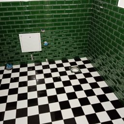 MK Remonty - Wykończenia - Świeżo wykafelkowana łazienka z błyszczącymi, szmaragdowozielonymi płytkami typu metro na ścianach i podłogą w czarno-białą szachownicę, widoczne przyłącza wodne i rewizja.