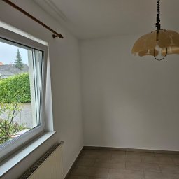 MK Remonty - Wykończenia - Pusty pokój z oknem wychodzącym na zieleń, wiszącą lampą w stylu retro i grzejnikiem pod oknem, przygotowany do malowania.