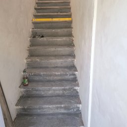 MK Remonty - Wykończenia - Surowe, betonowe schody w trakcie remontu, z poziomnicą na jednym ze stopni i butelką soku jabłkowego u podstawy, ściany pokryte świeżą gładzią.