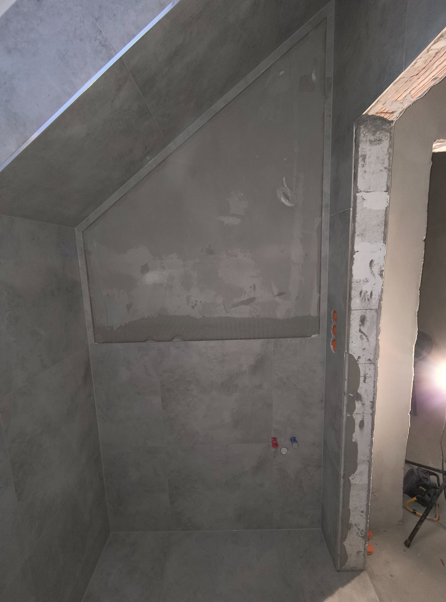 Szara łazienka w trakcie wykańczania, widoczne płytki imitujące beton, surowy beton na ścianie i przygotowane punkty instalacyjne.